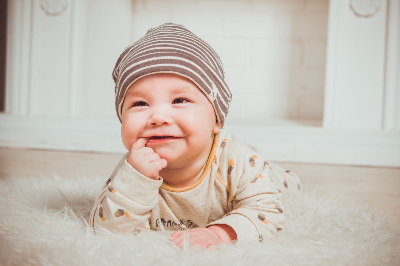 A smiling baby wearing a striped beanie and cozy outfit lies on a soft carpet indoors.